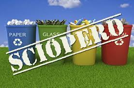 Quattro bidoni per la raccolta differenziata sono disposti all’aperto su un prato verde, con un cielo azzurro sullo sfondo. Da sinistra a destra i contenitori sono: blu per la carta, verde per il vetro, giallo per la plastica e rosso per i rifiuti non riciclabili. Ognuno è riempito con materiale corrispondente al tipo di raccolta. Sopra i bidoni, in diagonale e in grande evidenza, compare la scritta bianca “SCIOPERO”, come un timbro sovrapposto all’immagine.