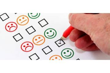 Mano che compila un questionario di soddisfazione segnando una casella accanto a una faccina sorridente verde, tra altre faccine con espressioni diverse.