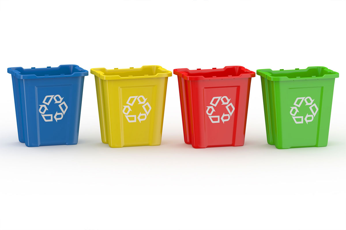 Immagine con quattro contenitori per la raccolta differenziata disposti in fila su sfondo chiaro. I bidoni sono di plastica e hanno tutti lo stesso design rettangolare con bordo superiore sporgente. Da sinistra a destra i colori sono: blu, giallo, rosso e verde. Su ciascun contenitore è presente il simbolo del riciclo bianco, ben visibile al centro. L’immagine è pulita, senza altri oggetti o elementi di distrazione, e comunica visivamente il concetto di separazione dei rifiuti.